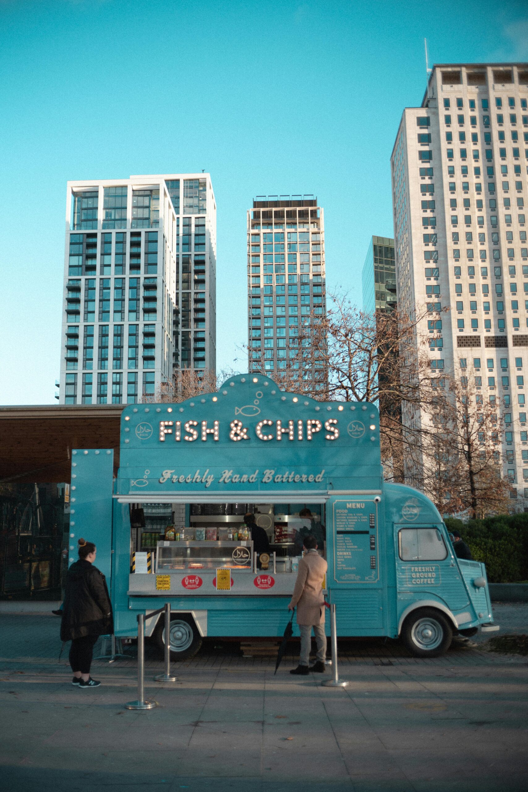 A vibrant blue food truck serves fish & chips amid urban skyscrapers, capturing urban life.